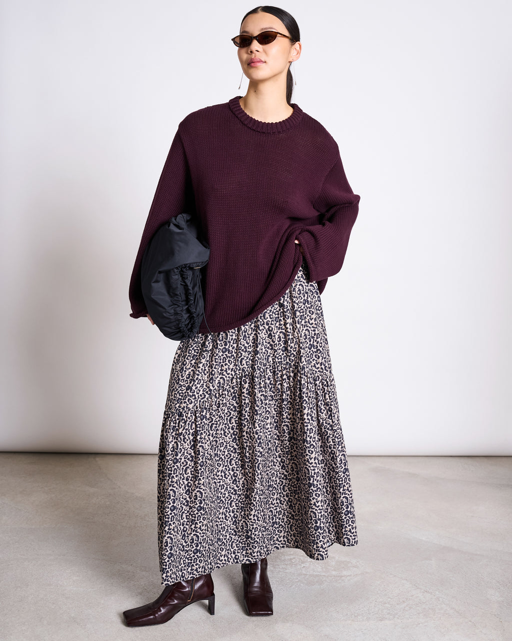 A woman wearing sunglasses, a loose maroon sweater, dark brown boots, and holding a black bag poses against a plain background in the jan-n-june TIERED MAXI SKIRT ALCA LEO PRINT.