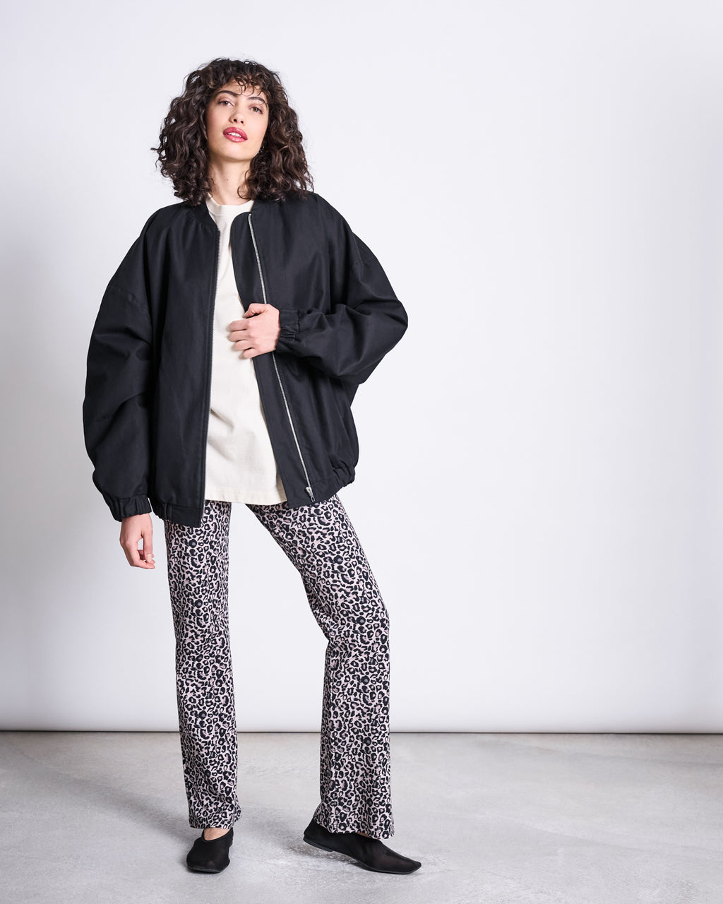 A woman with curly hair poses against a plain background, wearing jan-n-june’s FLARED LEGGINGS LEO GOTS, a loose black zip-up jacket, white shirt, and black shoes, with one hand on her hip as she looks to the side.