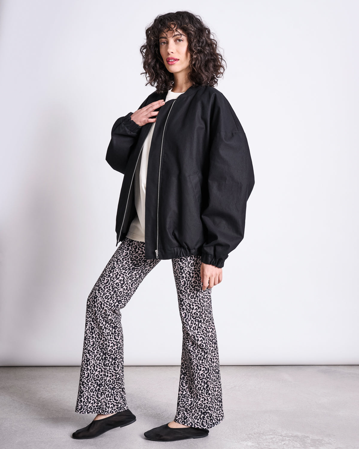 A person with curly hair poses against a plain background wearing jan-n-junes FLARED LEGGINGS LEO GOTS, a loose black jacket, white top, and black flats. They look at the camera in a relaxed pose.