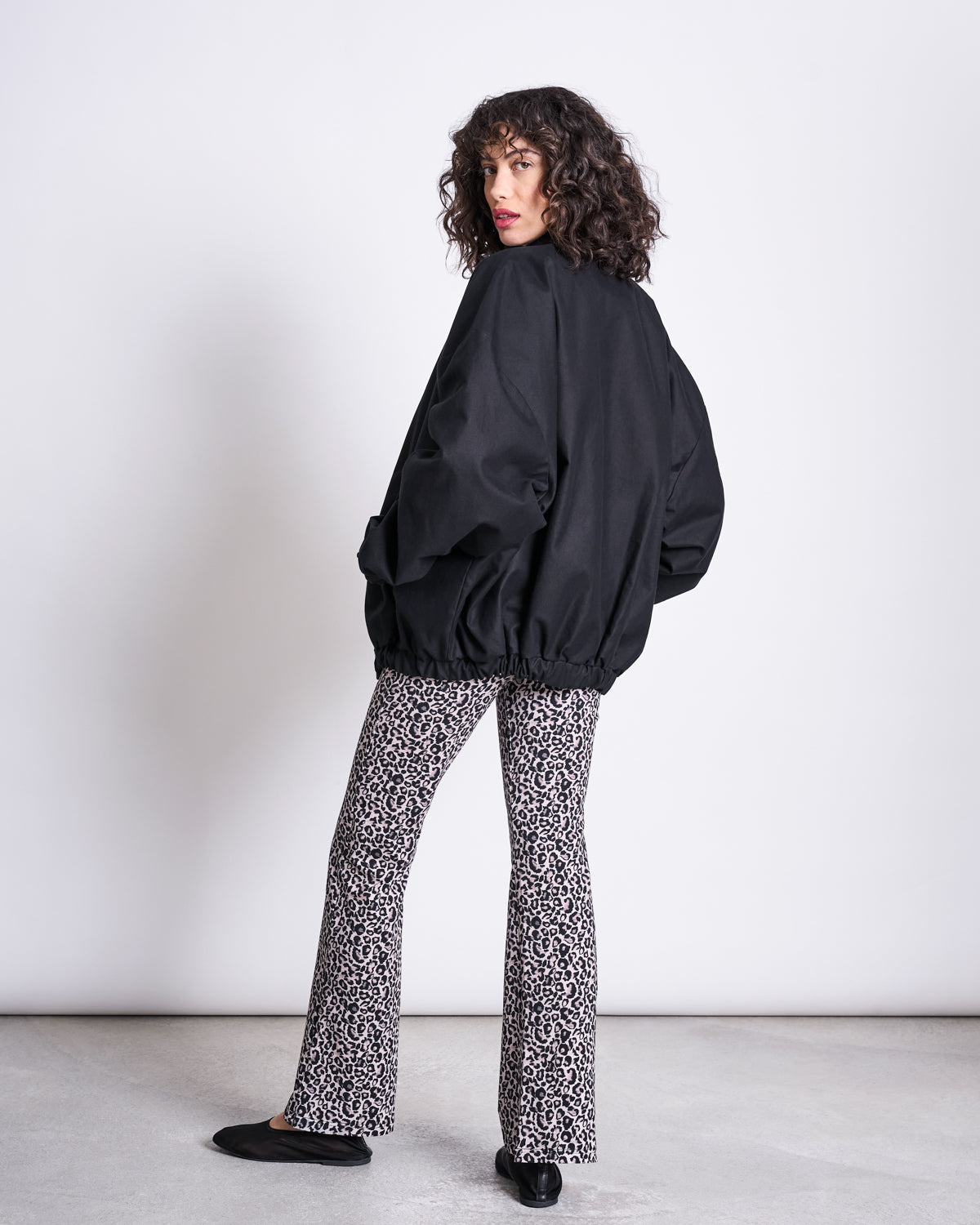 A person with curly hair, dressed in a loose black jacket, jan-n-june FLARED LEGGINGS LEO GOTS, and black shoes stands sideways against a plain light background.
