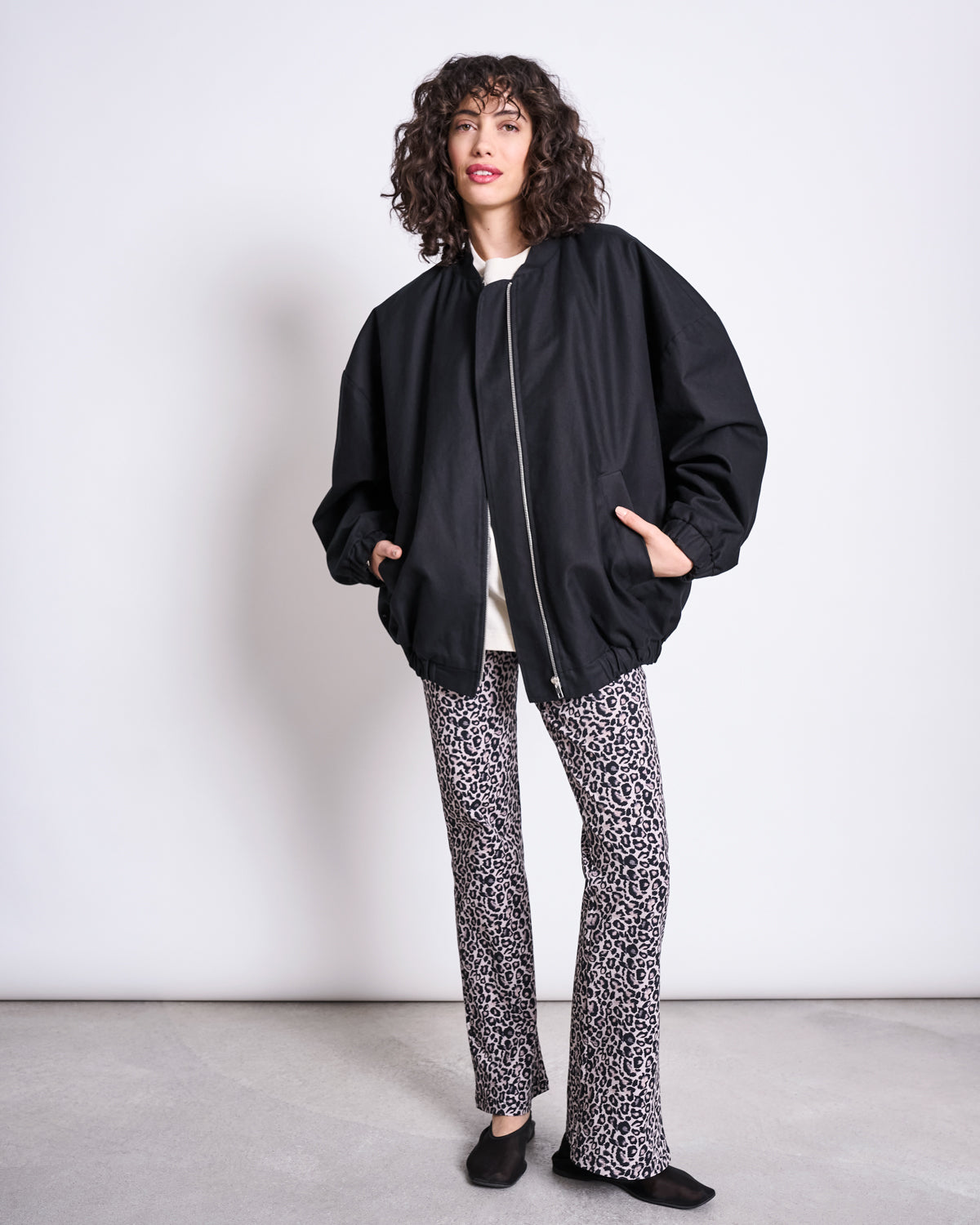 A person with curly hair wears an oversized black zip-up jacket, white top, jan-n-june’s FLARED LEGGINGS LEO GOTS, and black shoes, standing against a plain white wall and smiling slightly.