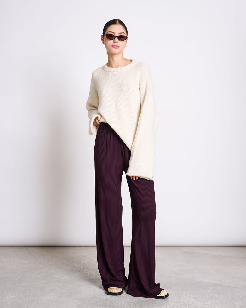 A person in sunglasses, an oversized cream sweater, jan-n-junes LYOCELL RIB PANTS SMILLA CHERRY LACQUER, and open-toe sandals stands against a plain white wall on a concrete floor.