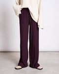 A person wearing jan-n-june’s LYOCELL RIB PANTS SMILLA CHERRY LACQUER, paired with a cream oversized sweater and beige sandals, stands on a light gray floor against a plain white background.