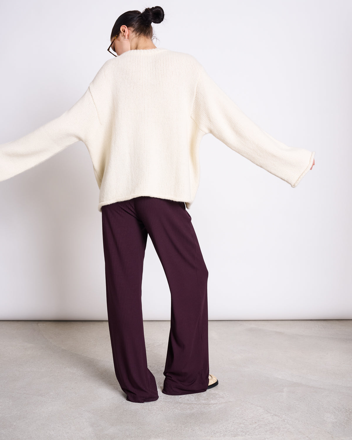 A person stands with arms outstretched, wearing jan-n-june’s LYOCELL RIB PANTS SMILLA CHERRY LACQUER—wide-legged, eco-friendly pants in soft Tencel™—and a loose cream sweater. A plain background and concrete floor enhance the minimal look.
