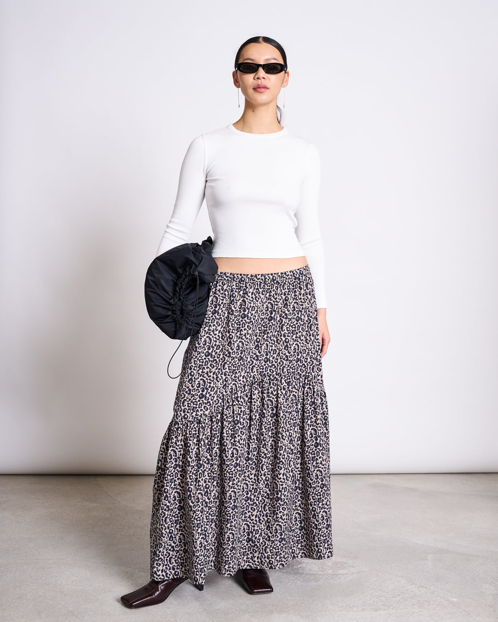 A woman in sunglasses wears a fitted white long-sleeve top and the jan-n-june TIERED MAXI SKIRT ALCA LEO PRINT, made from 100% cotton. She holds a black ruched bag, standing on a concrete floor against a plain white background.