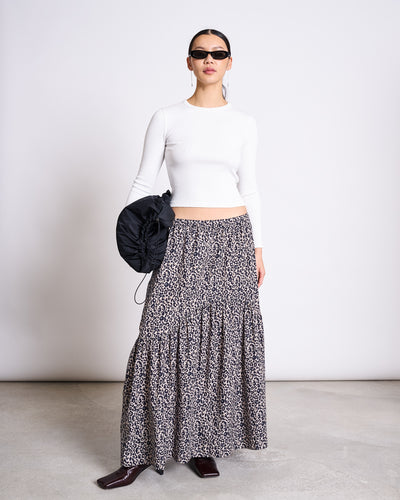 A woman in sunglasses wears a fitted white long-sleeve top and the jan-n-june TIERED MAXI SKIRT ALCA LEO PRINT, made from 100% cotton. She holds a black ruched bag, standing on a concrete floor against a plain white background.