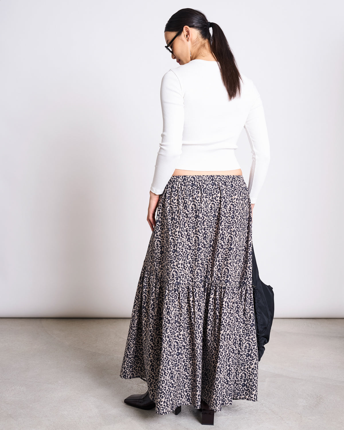 A woman with dark hair in a ponytail stands with her back to the camera, wearing the jan-n-june RIB LONGSLEEVE MURI WHITE GOTS top paired with a long, tiered leopard-print skirt, black shoes, and glasses—a perfect layering piece.
