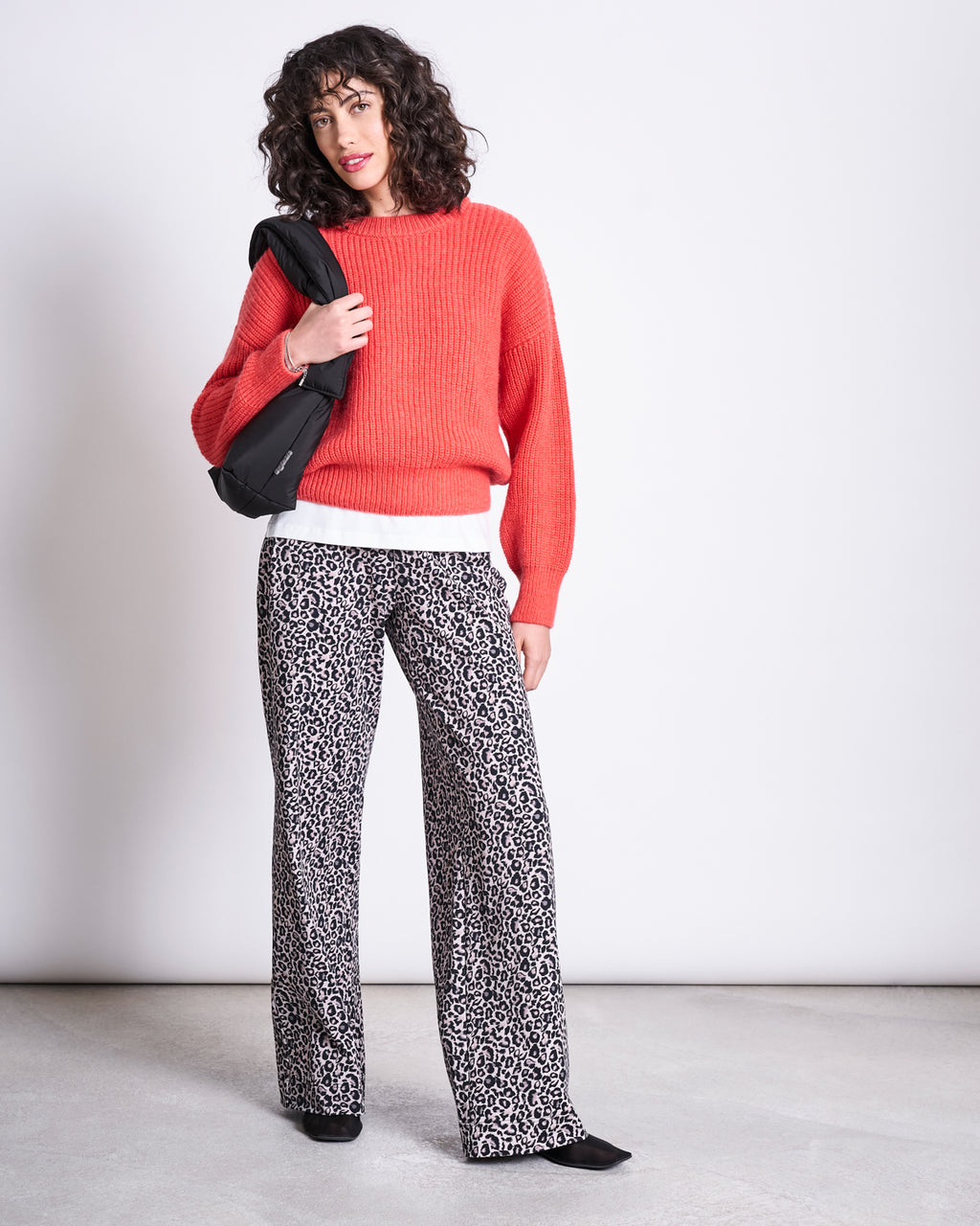 A woman with curly hair models jan-n-junes PANTS KISO LEO GOTS, paired with a red sweater, white top, black shoes, and a black shoulder bag, standing against a plain white background.