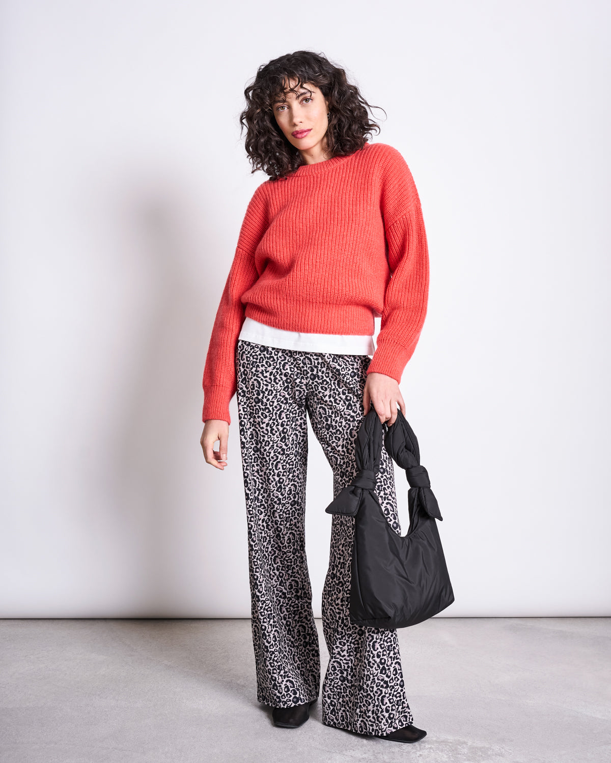 A person with curly hair wears a red sweater, white shirt, and jan-n-june PANTS KISO LEO GOTS, holding a black handbag while standing against a plain light-colored background.
