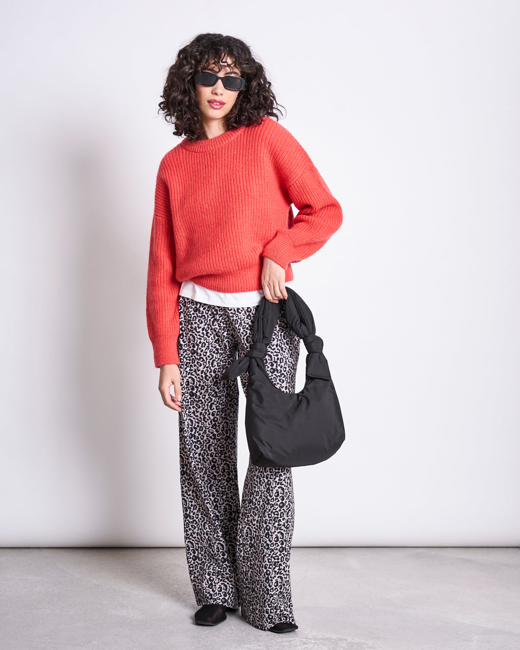 A person stands against a plain white wall, wearing a bright red sweater, white shirt, leopard print wide-leg pants, black shoes, sunglasses, and holding the jan-n-june BAG BIWA PUFFY MINI BLACK.