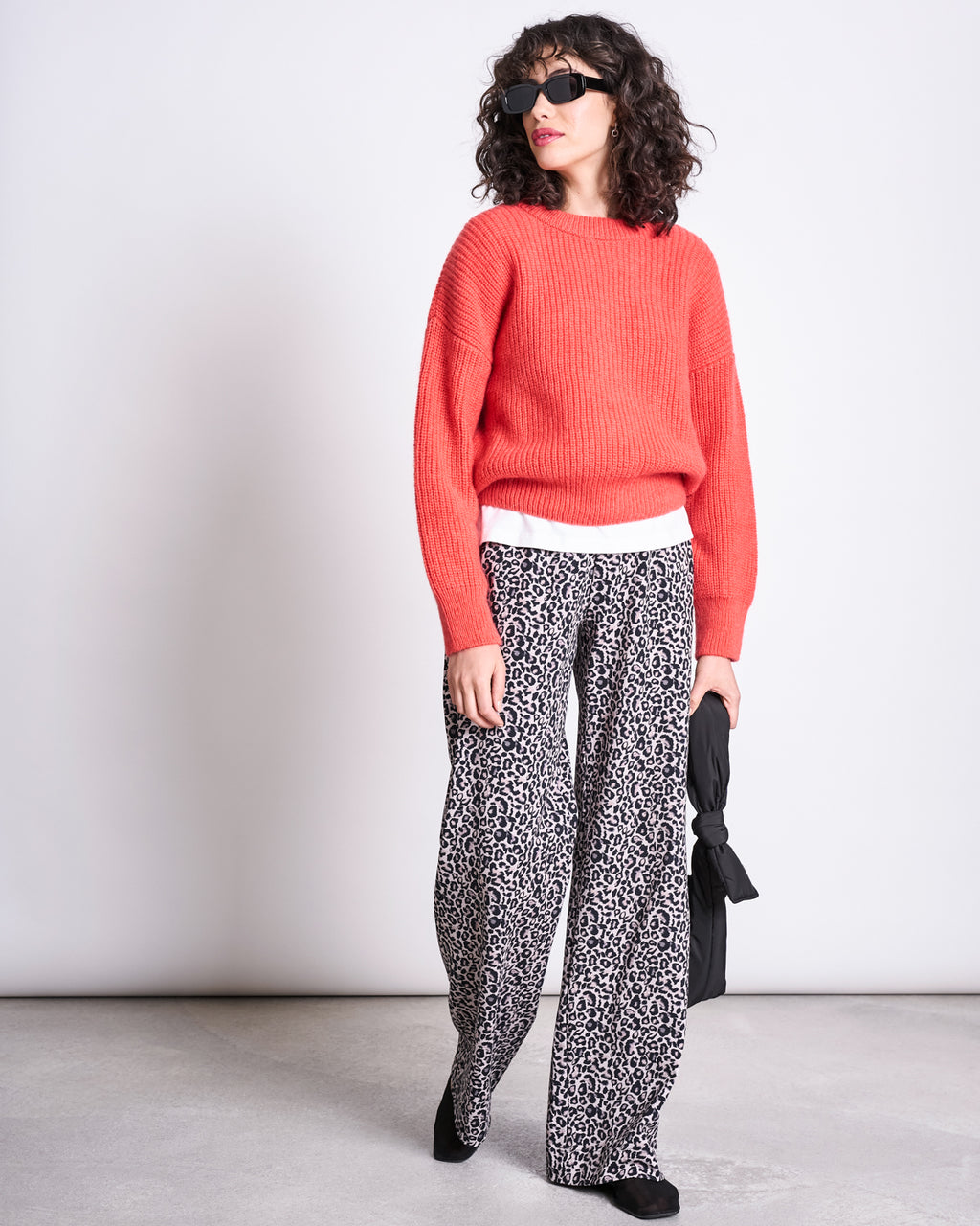 A woman in jan-n-junes PANTS KISO LEO GOTS, a bright red sweater, white undershirt, black shoes, and sunglasses stands against a plain background holding a folded black umbrella by her side.