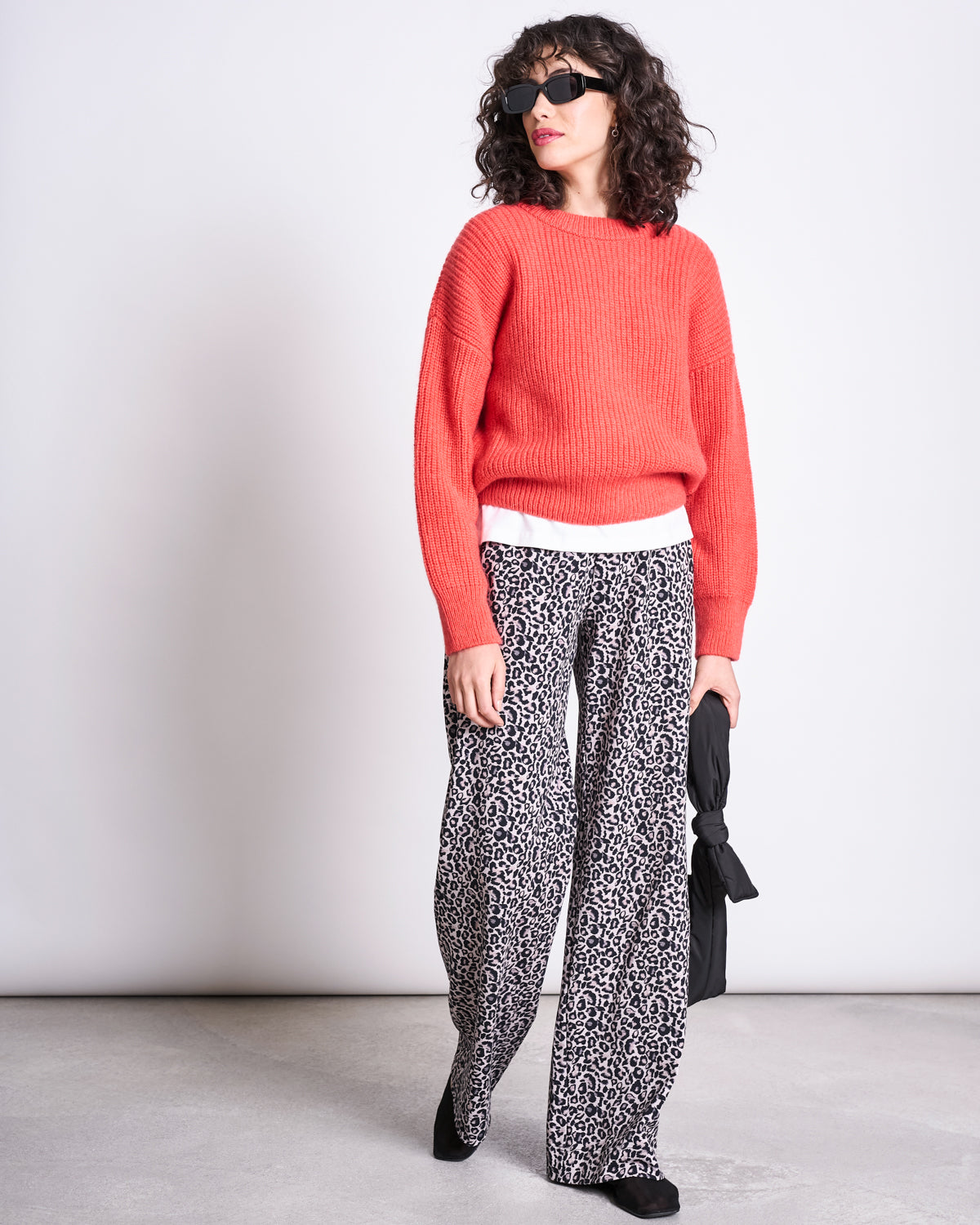 A woman in jan-n-junes PANTS KISO LEO GOTS, a bright red sweater, white undershirt, black shoes, and sunglasses stands against a plain background holding a folded black umbrella by her side.