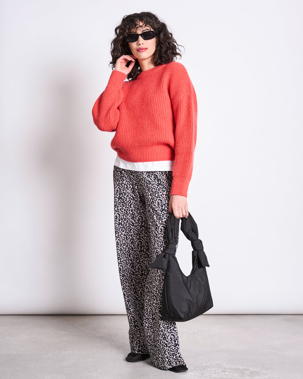 A woman wearing sunglasses, a bright red-orange sweater, and jan-n-june PANTS KISO LEO GOTS stands against a plain background, holding a black bag with knotted handles.