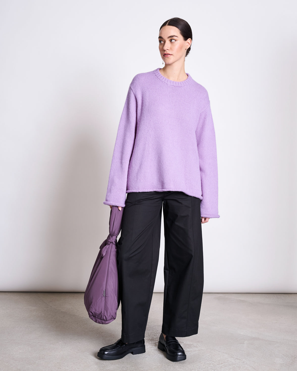 A woman poses against a plain backdrop in the jan-n-june OVERSIZED ALPACA MIX JUMPER WOOLY SONA LAVENDER, paired with black wide-leg pants, black shoes, and holding a purple bag. She looks slightly right with a neutral expression.