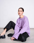 A woman with dark hair tied back sits on the floor against a white background, wearing the jan-n-june OVERSIZED ALPACA MIX JUMPER WOOLY SONA LAVENDER, black wide-leg pants, and black loafers, looking neutrally at the camera.