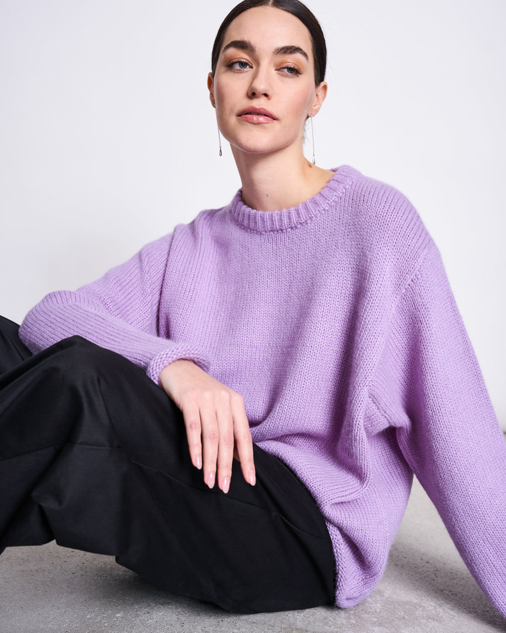 A woman with dark hair pulled back wears the jan-n-june OVERSIZED ALPACA MIX JUMPER WOOLY SONA LAVENDER and black pants, sitting on the floor with one arm on her knee, confidently looking to the side against a plain background.