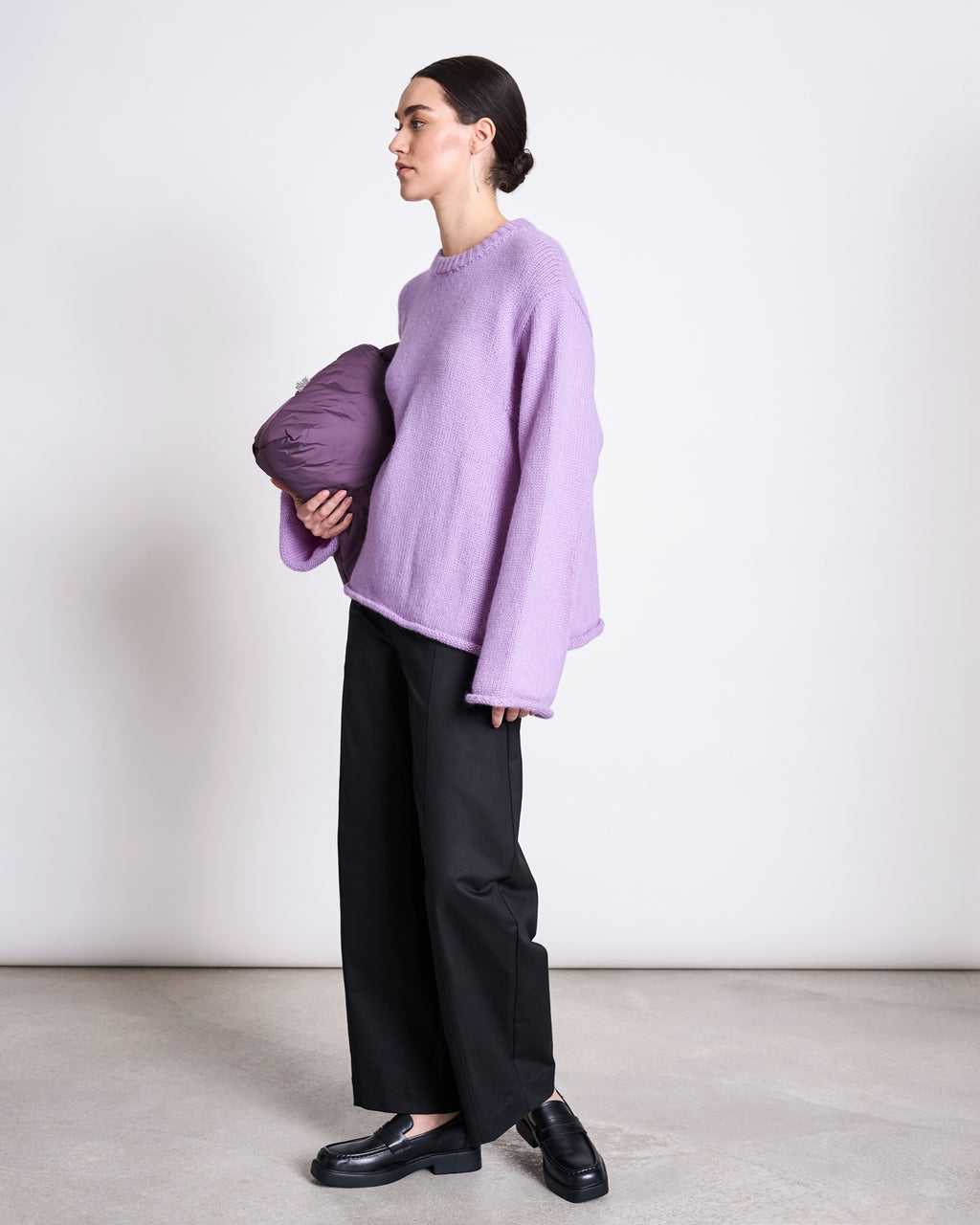 A person stands against a plain background, wearing a loose lavender sweater, black loafers, and jan-n-june ORGANIC COTTON BARREL PANTS YAURI BLACK, holding a purple puffy item under one arm.
