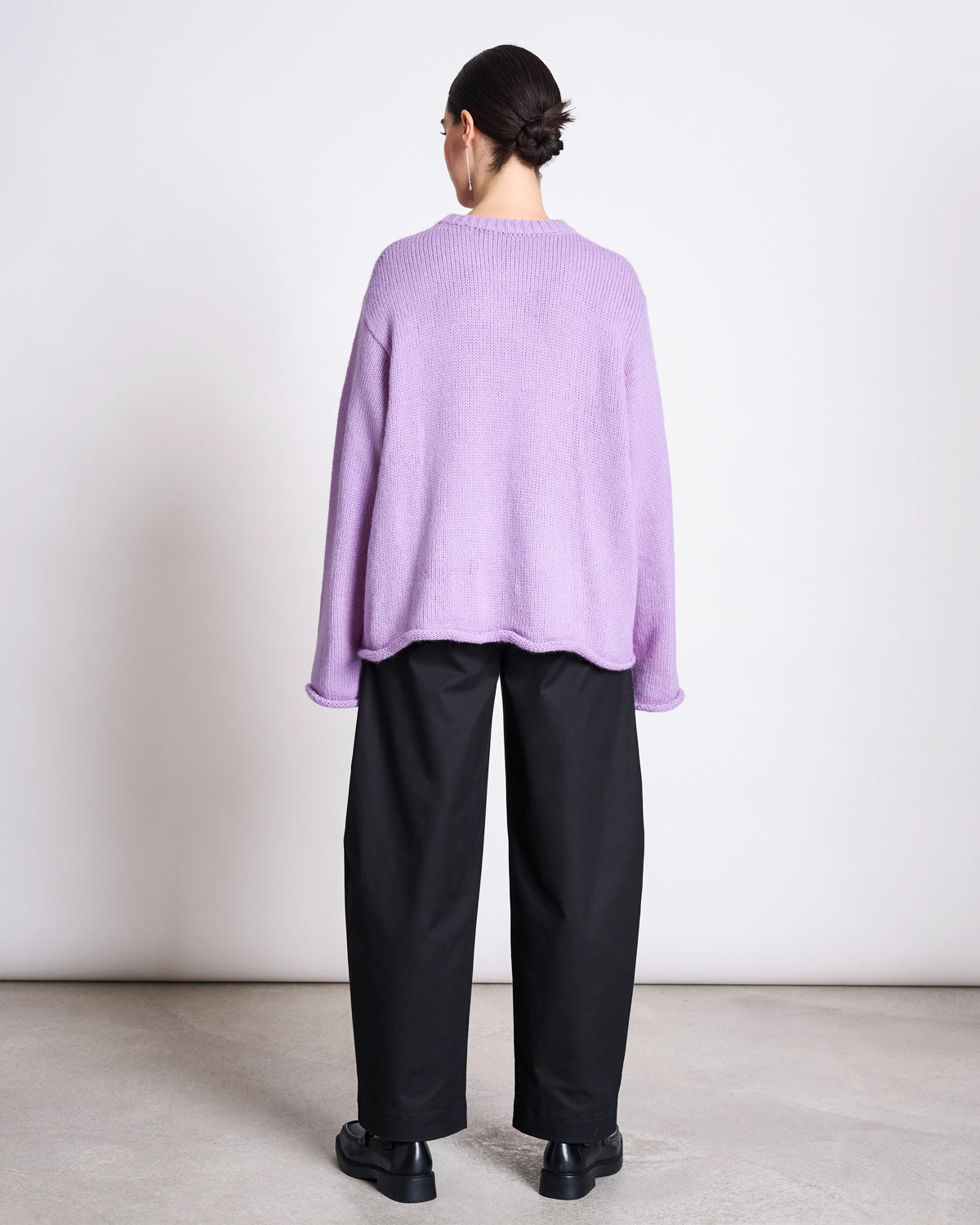 A person with a low bun stands facing away from the camera, wearing a loose lavender sweater and jan-n-june's ORGANIC COTTON BARREL PANTS YAURI BLACK, paired with black shoes. The background is plain and light-colored.
