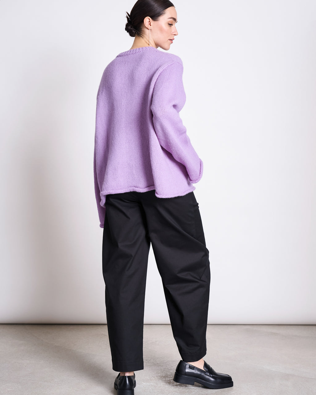 A person stands sideways, glancing over their shoulder in a loose lavender sweater and jan-n-june's ORGANIC COTTON BARREL PANTS YAURI BLACK, paired with black loafers. Their dark hair is pulled back against a plain, light background.