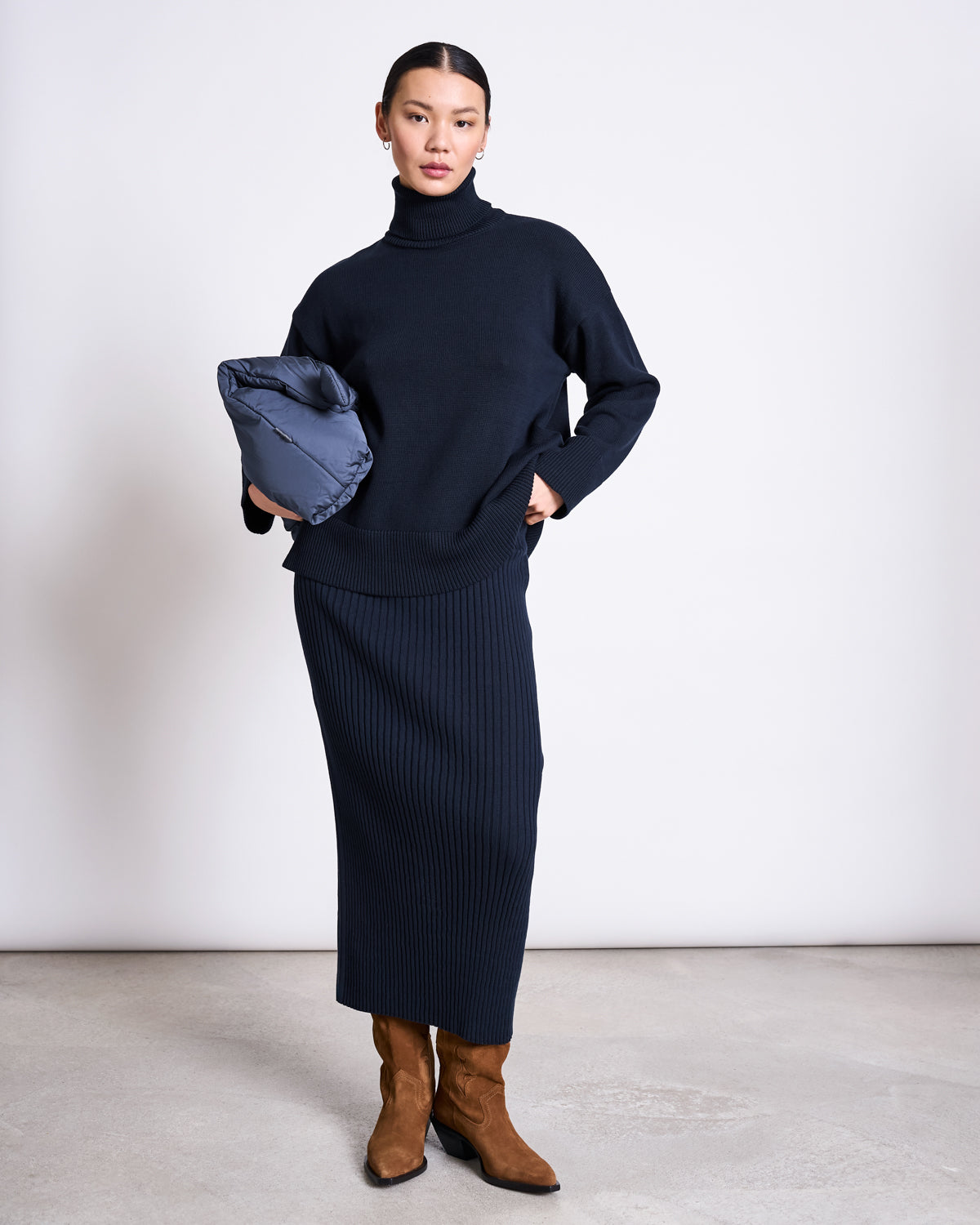 A woman with her hair pulled back stands against a plain background, wearing jan-n-junes TURTLENECK MUA NIGHTFALL GOTS sweater, a long ribbed black skirt, and brown suede boots, holding a folded gray jacket in her left arm.