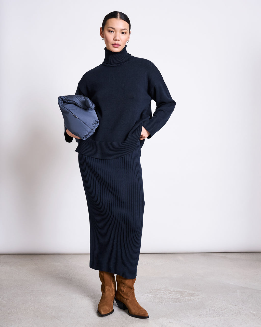 A woman wearing the jan-n-june MIDI KNIT SKIRT EBBA NIGHTFALL GOTS, a black turtleneck sweater, and brown boots stands against a plain background, holding a folded blue jacket under her arm.
