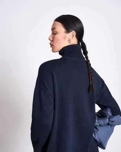A woman with a long dark braid wears the jan-n-june TURTLENECK MUA NIGHTFALL GOTS, made from 100% organic cotton, standing sideways and looking down with her arm behind her against a plain light background.