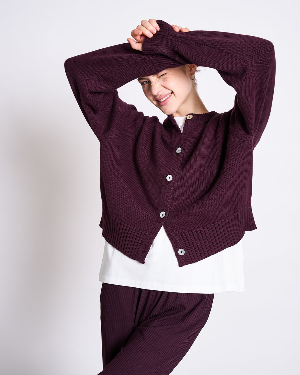 A person smiles playfully with arms raised, wearing the jan-n-june RELAXED CREWNECK CARDIGAN SIVIA CHERRY LACQUER GOTS and matching pants over a white shirt. Made from GOTS certified materials for added comfort.