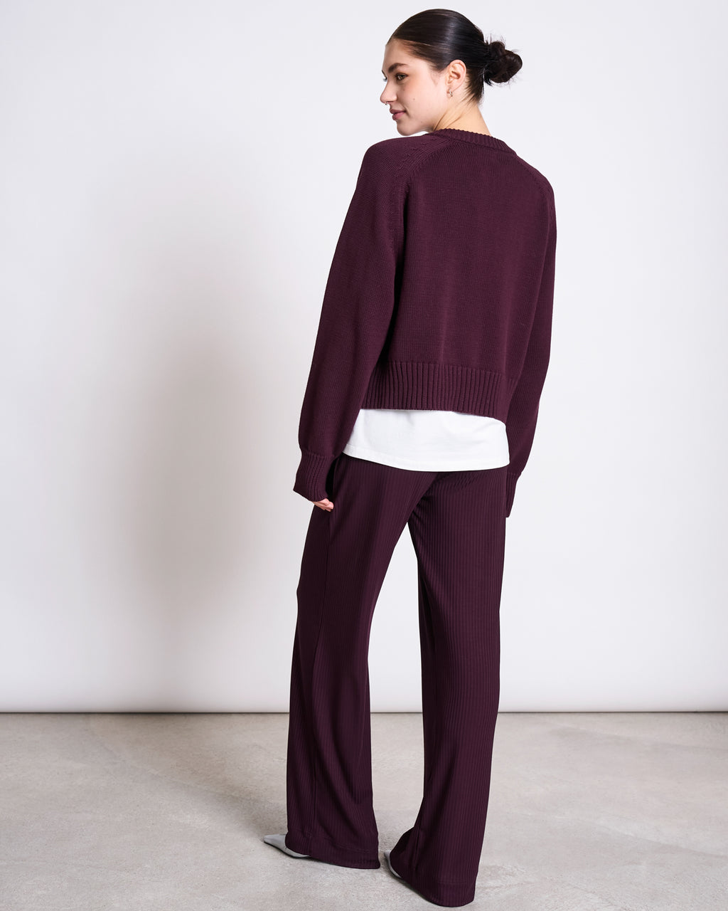 A woman with a neat bun faces away, wearing the jan-n-june RELAXED CREWNECK CARDIGAN SIVIA in Cherry Lacquer over a white shirt and matching burgundy ribbed sweater and pants, set against a plain light background.