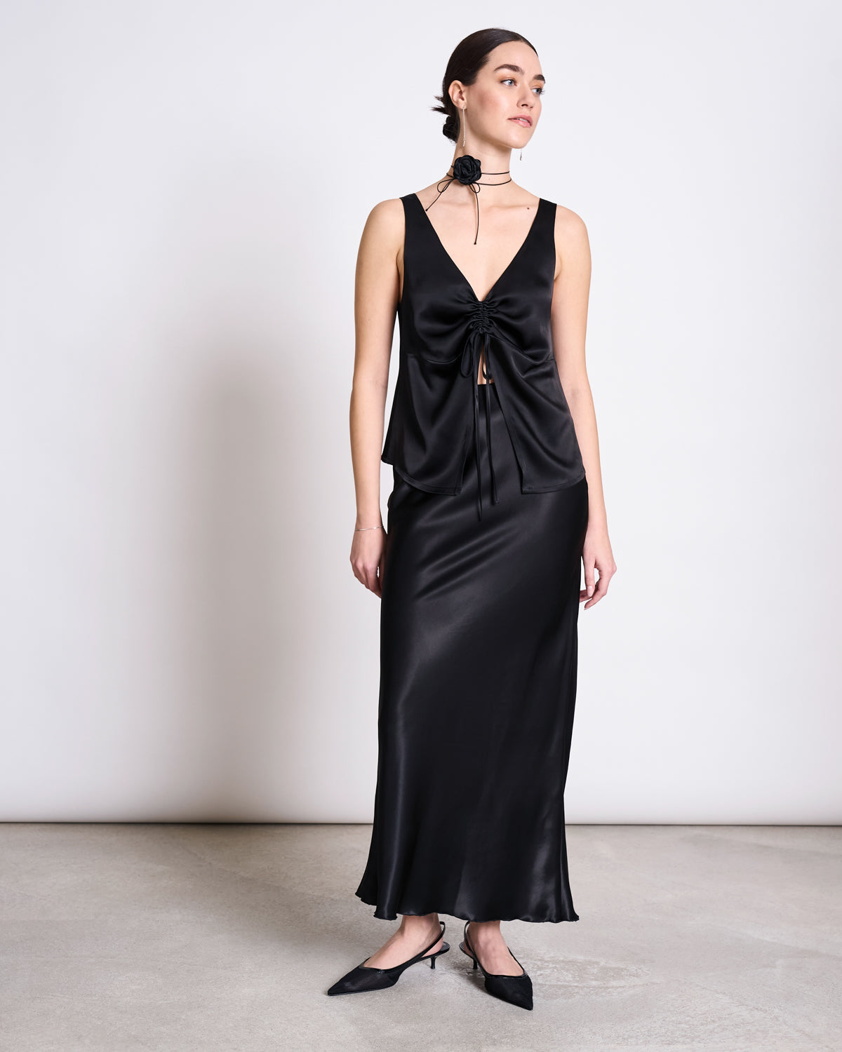 A woman poses in a minimalist setting wearing the jan-n-june RUFFLED V-NECK TOP TALARA BLACK with a matching long satin skirt, styled with a floral black choker and pointed black heels.