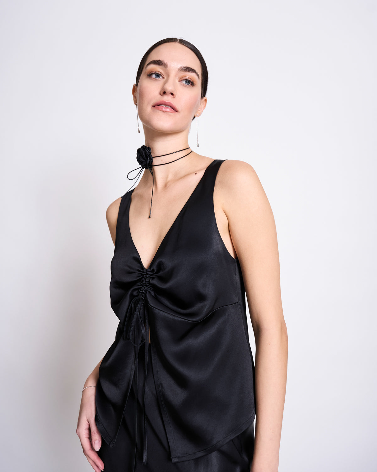 A woman with dark hair tied back wears the sleeveless jan-n-june RUFFLED V-NECK TOP TALARA BLACK, featuring a deep V-neck and gathered drawstring detail, paired with a black flower choker, standing against a plain light background.