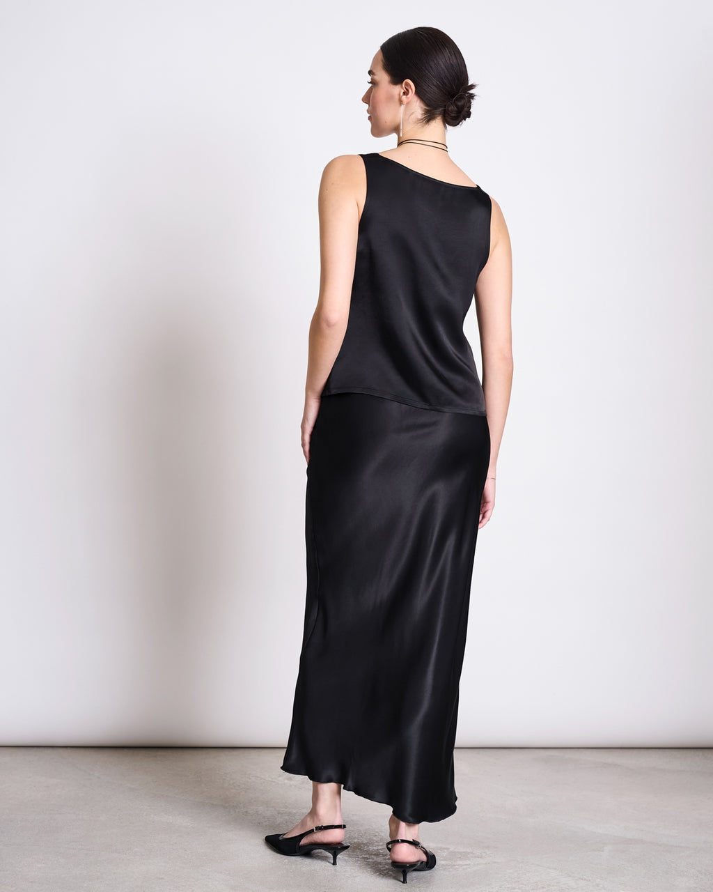 A woman with dark hair in a low bun stands facing away from the camera, wearing the jan-n-june RUFFLED V-NECK TOP TALARA BLACK paired with a matching long skirt and black sandals against a plain light background.