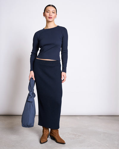 A person with hair pulled back and a neutral expression stands indoors against a light wall, wearing the jan-n-june MIDI KNIT SKIRT EBBA NIGHTFALL GOTS, a matching navy long-sleeve top, brown boots, and holding a rolled-up navy jacket.
