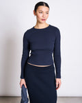 A woman wears the jan-n-june RIB LONGSLEEVE MURI NIGHTFALL GOTS, a slim-fitting navy top, with a matching ribbed skirt. She stands against a plain light background, holding a navy item in her left hand and looking slightly to the side.
