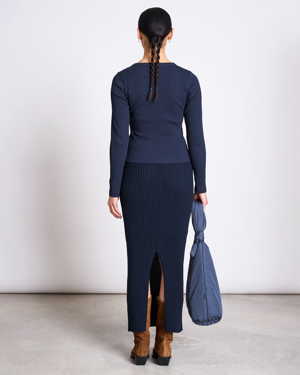 A woman with two braids faces away, wearing the jan-n-june RIB LONGSLEEVE MURI NIGHTFALL GOTS top and a ribbed navy skirt with a back slit, paired with brown boots and a blue quilted bag. The background is plain and neutral.