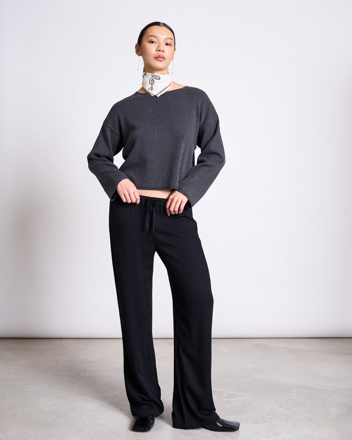 A woman stands against a plain background in a capsule wardrobe look: the jan-n-june LIGHT KNIT JUMPER WILLIAMSBURG ANTHRACITE GOTS, loose black pants, black shoes, and a white subtly patterned scarf around her neck.