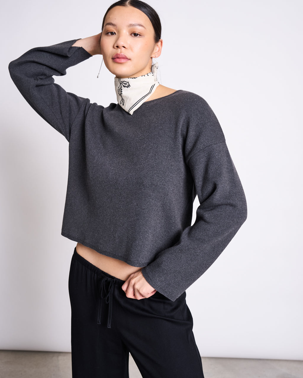 A woman poses with one hand behind her head, wearing the jan-n-june LIGHT KNIT JUMPER WILLIAMSBURG ANTHRACITE GOTS—an organic cotton long-sleeve top—styled with black pants and a white patterned scarf against a plain light background.