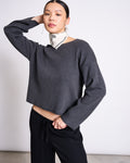 A woman poses with one hand behind her head, wearing the jan-n-june LIGHT KNIT JUMPER WILLIAMSBURG ANTHRACITE GOTS—an organic cotton long-sleeve top—styled with black pants and a white patterned scarf against a plain light background.