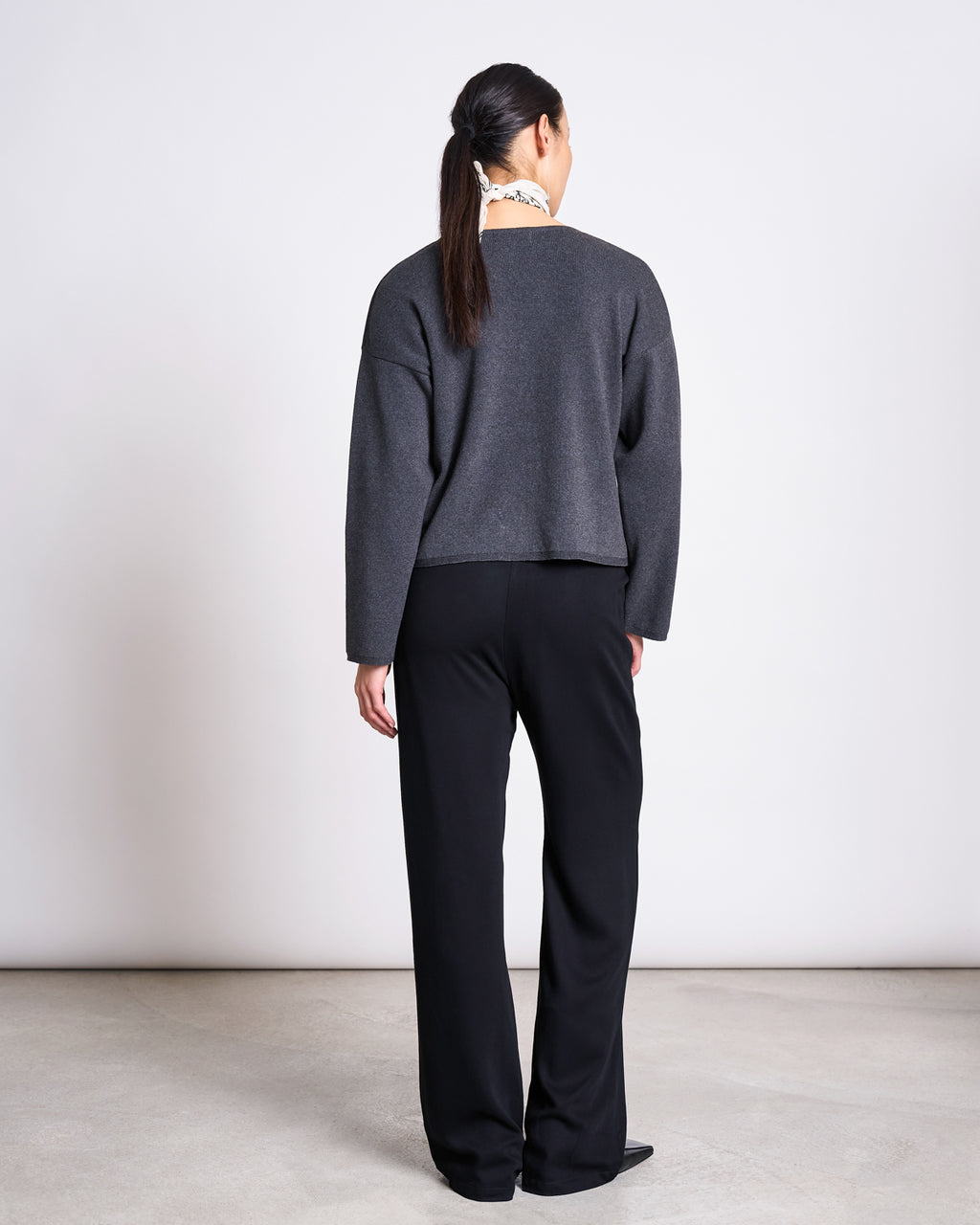 A person with long dark hair in a ponytail stands facing away, wearing the jan-n-june LIGHT KNIT JUMPER WILLIAMSBURG ANTHRACITE GOTS and black wide-leg pants—timeless staples—against a light wall and concrete floor.