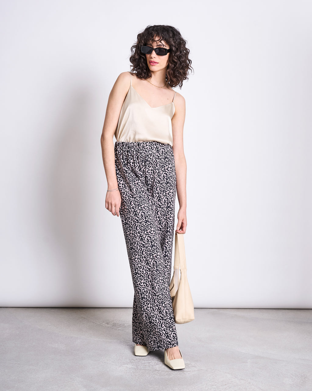 A woman with curly hair wearing sunglasses, a beige camisole, jan-n-june PANTS KISO LEO GOTS, cream square-toe shoes, and holding a beige bag poses against a plain light background.