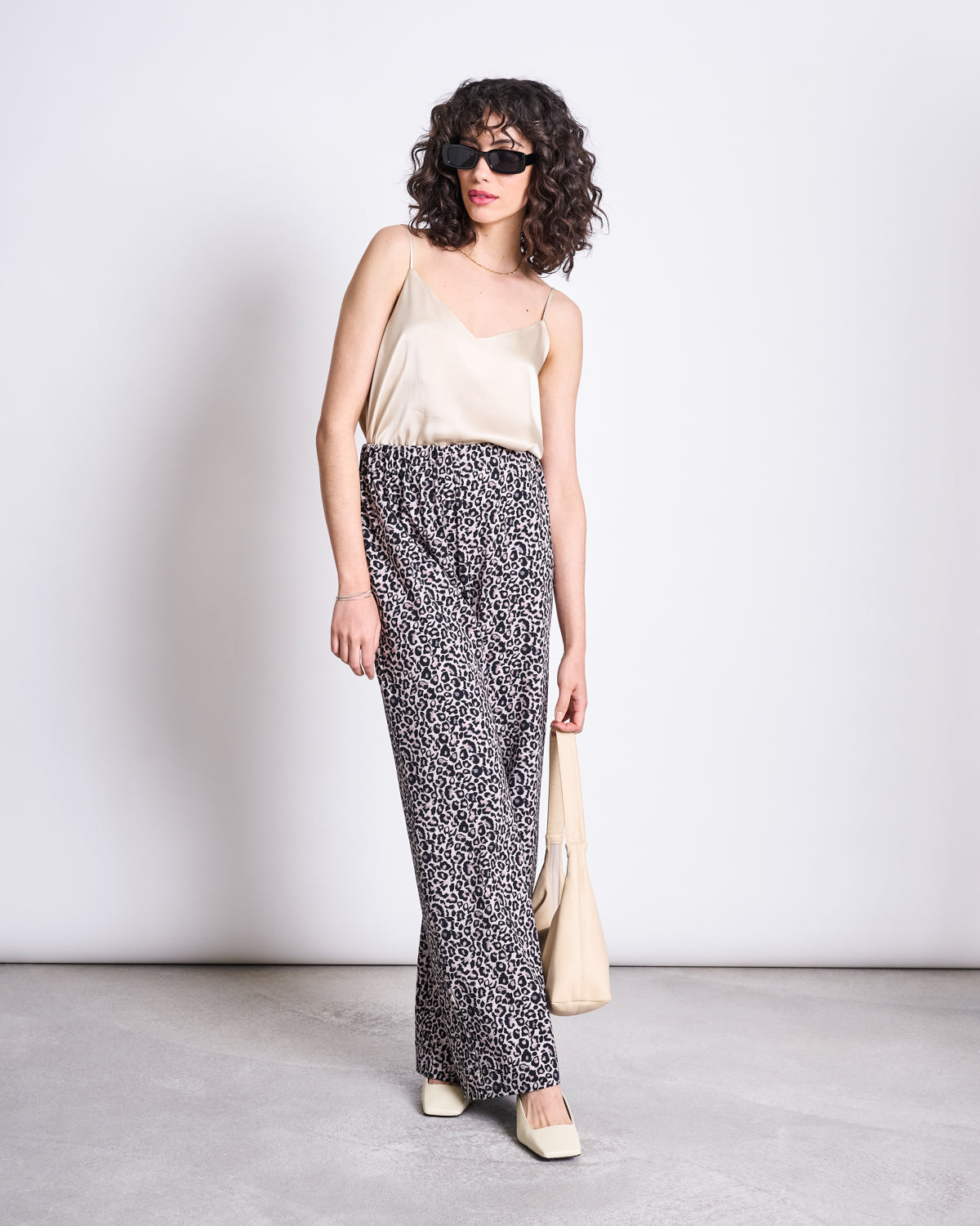 A woman with curly hair wearing sunglasses, a beige camisole, jan-n-june PANTS KISO LEO GOTS, cream square-toe shoes, and holding a beige bag poses against a plain light background.