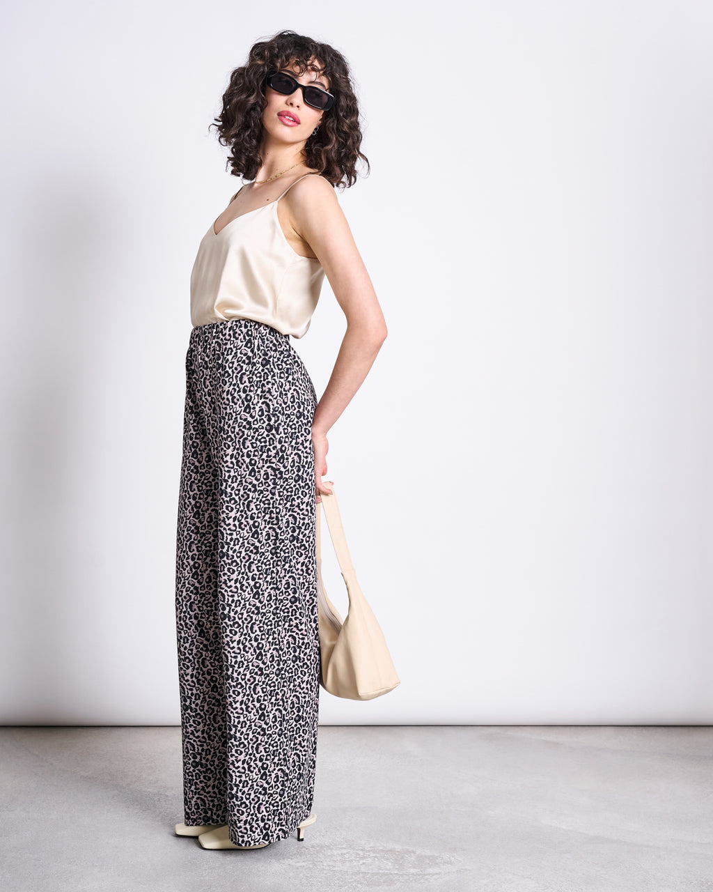 A woman in sunglasses wears jan-n-junes PANTS KISO LEO GOTS, a cream satin camisole, cream heels, and holds a matching cream handbag, standing against a plain light background.