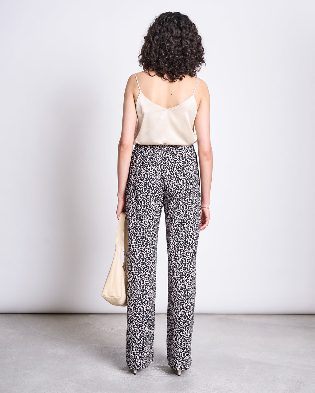 A woman with curly hair faces away, wearing jan-n-june’s PANTS KISO LEO GOTS—high-waisted leo print pants—with a cream satin camisole and handbag, standing against a white wall on a concrete floor.