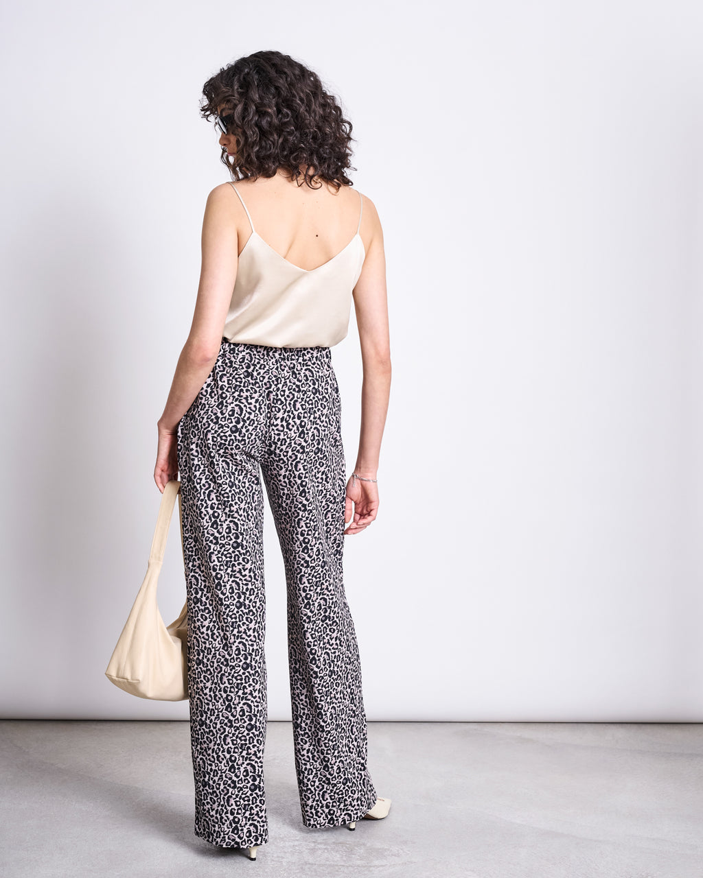 A woman with curly hair faces away in a minimal room, wearing the jan-n-june PANTS KISO LEO GOTS, a cream camisole, and holding a matching cream shoulder bag. The scene features a plain white wall and gray floor.