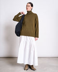 A woman stands against a plain background wearing the jan-n-june TURTLENECK MUA WOODY OLIVE GOTS sweater with a long white skirt, tan boots, and a black shoulder bag. Her hair is pulled back neatly.