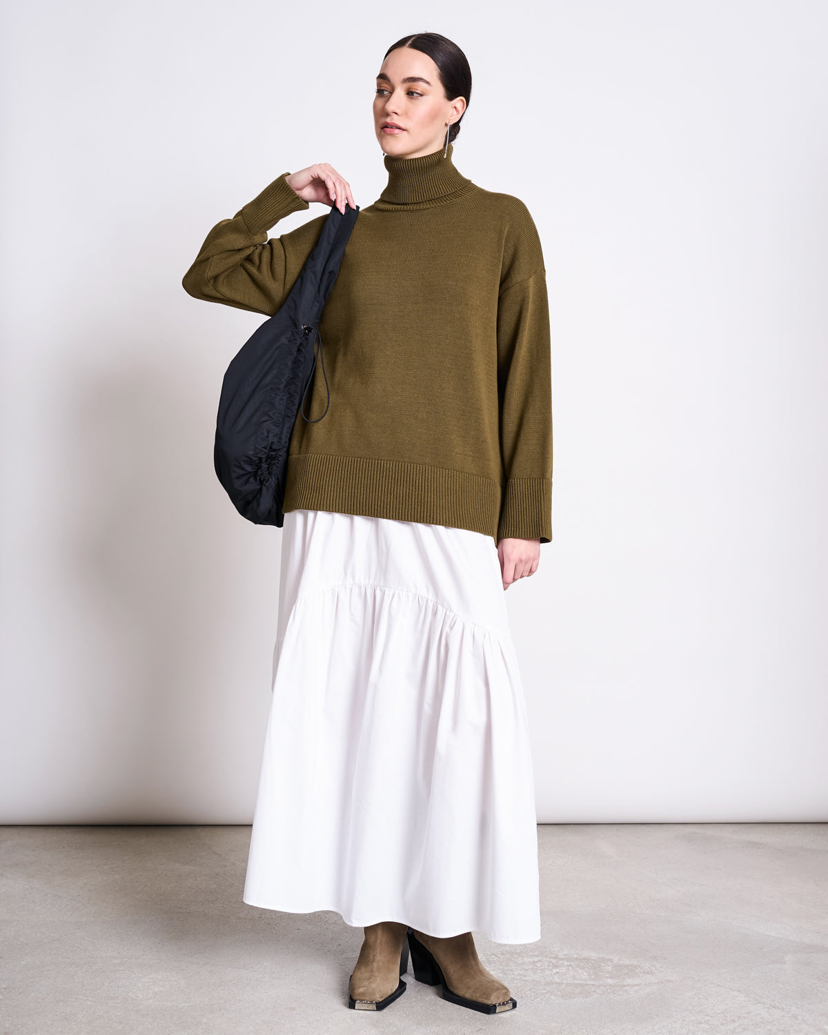 A woman stands against a plain background wearing the jan-n-june TURTLENECK MUA WOODY OLIVE GOTS sweater with a long white skirt, tan boots, and a black shoulder bag. Her hair is pulled back neatly.