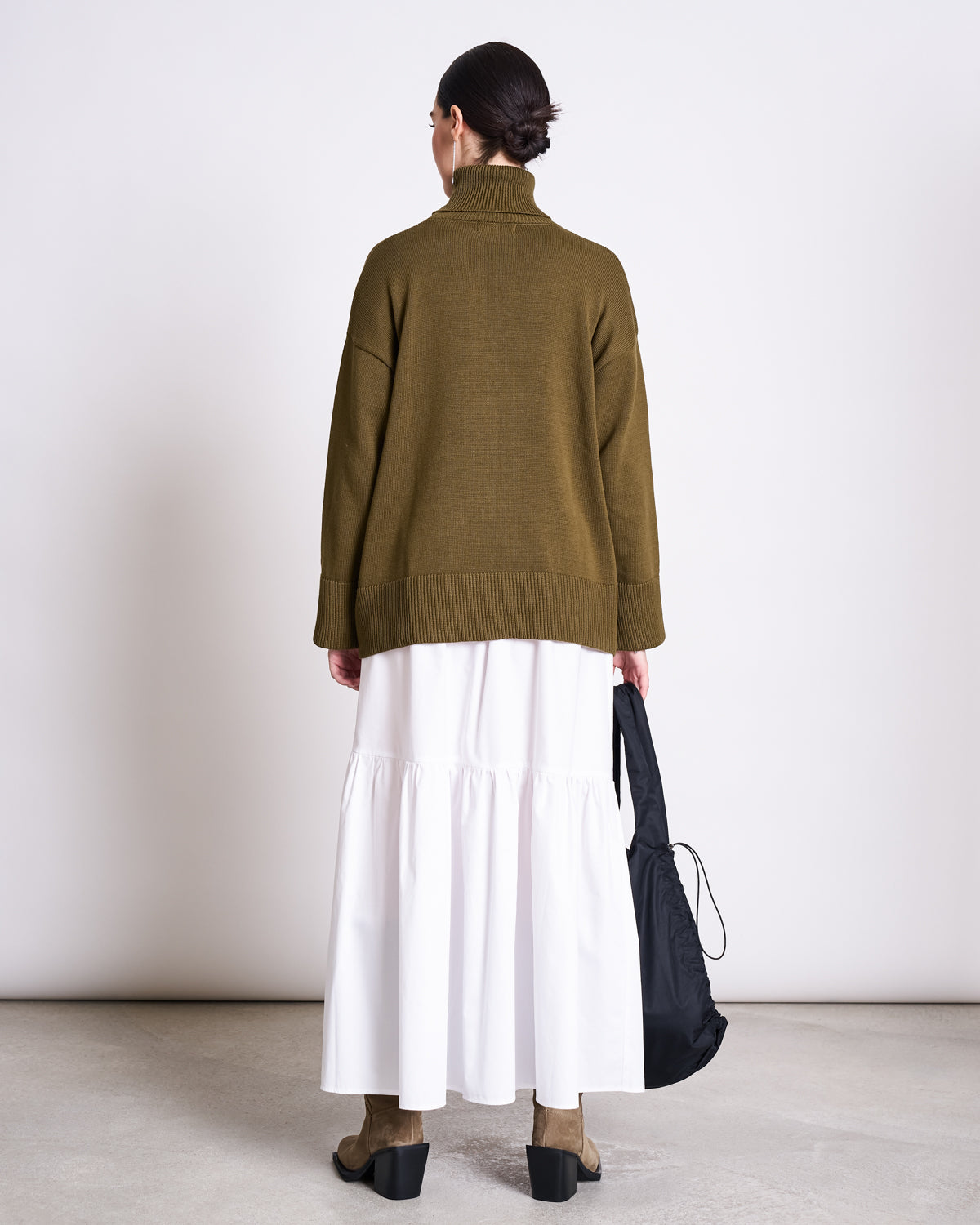 A person stands with their back to the camera, wearing the jan-n-june TURTLENECK MUA WOODY OLIVE GOTS sweater, a long white skirt, taupe boots, and holding a black bag against a plain light background.