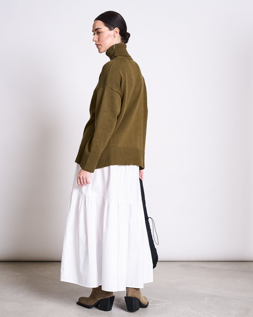 A woman stands sideways against a plain white background wearing the jan-n-june TURTLENECK MUA WOODY OLIVE GOTS, paired with a long white tiered skirt, taupe ankle boots, and holding a black bag.