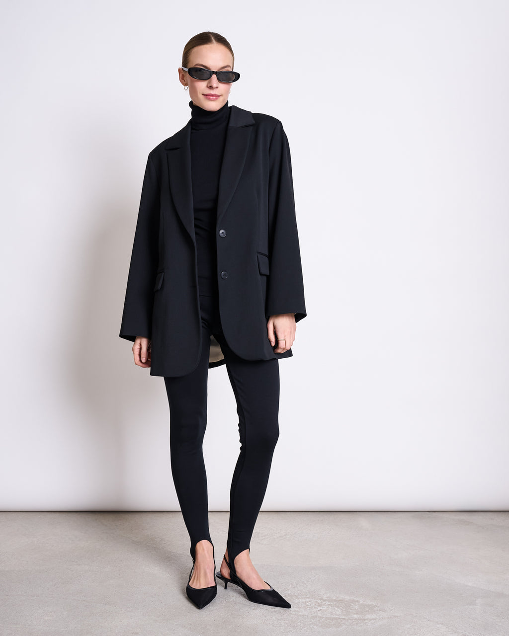 A woman stands against a plain wall wearing black sunglasses, an oversized blazer, a black turtleneck, jan-n-junes STIRRUP LEGGINGS BLACK, and pointed black flats. Her hair is pulled back and her hands rest by her sides.