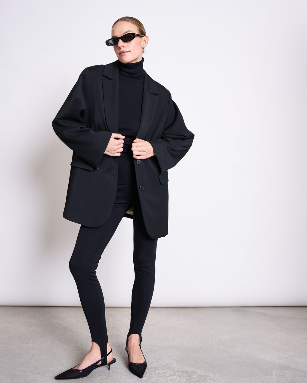 A person stands against a plain white wall wearing black sunglasses, a black turtleneck, a loose black blazer, jan-n-june STIRRUP LEGGINGS BLACK, and black pointed-toe slingback heels.