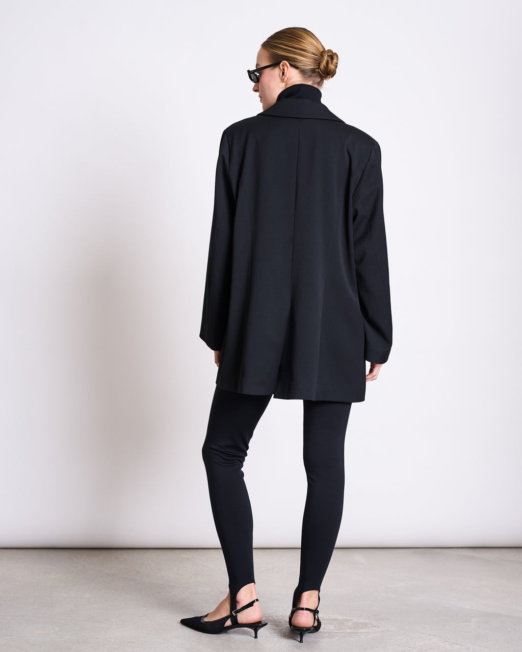 A woman with a neat low bun, wearing jan-n-june STIRRUP LEGGINGS BLACK, a black blazer, pointed black flats, and sunglasses stands facing away against a plain light background.